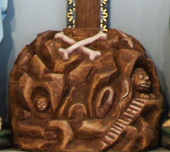 Notice the “Golgotha base” on which the crucifix is planted. The skull and crossbones are symbols of death. Showing them under Jesus’ feet symbolises Christ conquering Death by His death. Among the Jews it was traditionally represented that the skull of Adam, after being handed by Noah to his son Shem, and by him to Melchisedech, was finally deposited at Golgotha, which in Aramaic means ‘the place of the skull’. It was probably a small hill just outside the walls of ancient Jerusalem. Curiously the Golgotha base here depicts not one but two skulls.